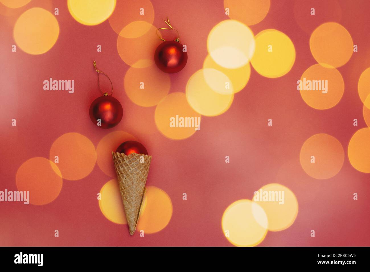 Weihnachtskugeln in einem Waffelkegel auf rotem Hintergrund und orangefarbenem hellem Bokeh. Runde Spielzeuge mit Goldfaden. Alternatives Konzept der Feier von Winterferien, Draufsicht. Flach Liegend Stockfoto