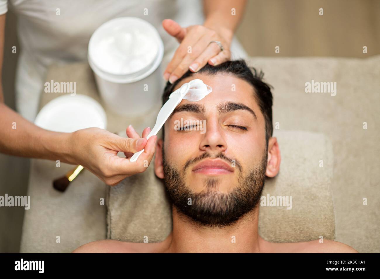 Draufsicht der anonymen Frau, die während der Arbeit im Schönheitssalon eine reinigende Maske auf das Gesicht des bärtigen jungen männlichen Klienten anwendet Stockfoto