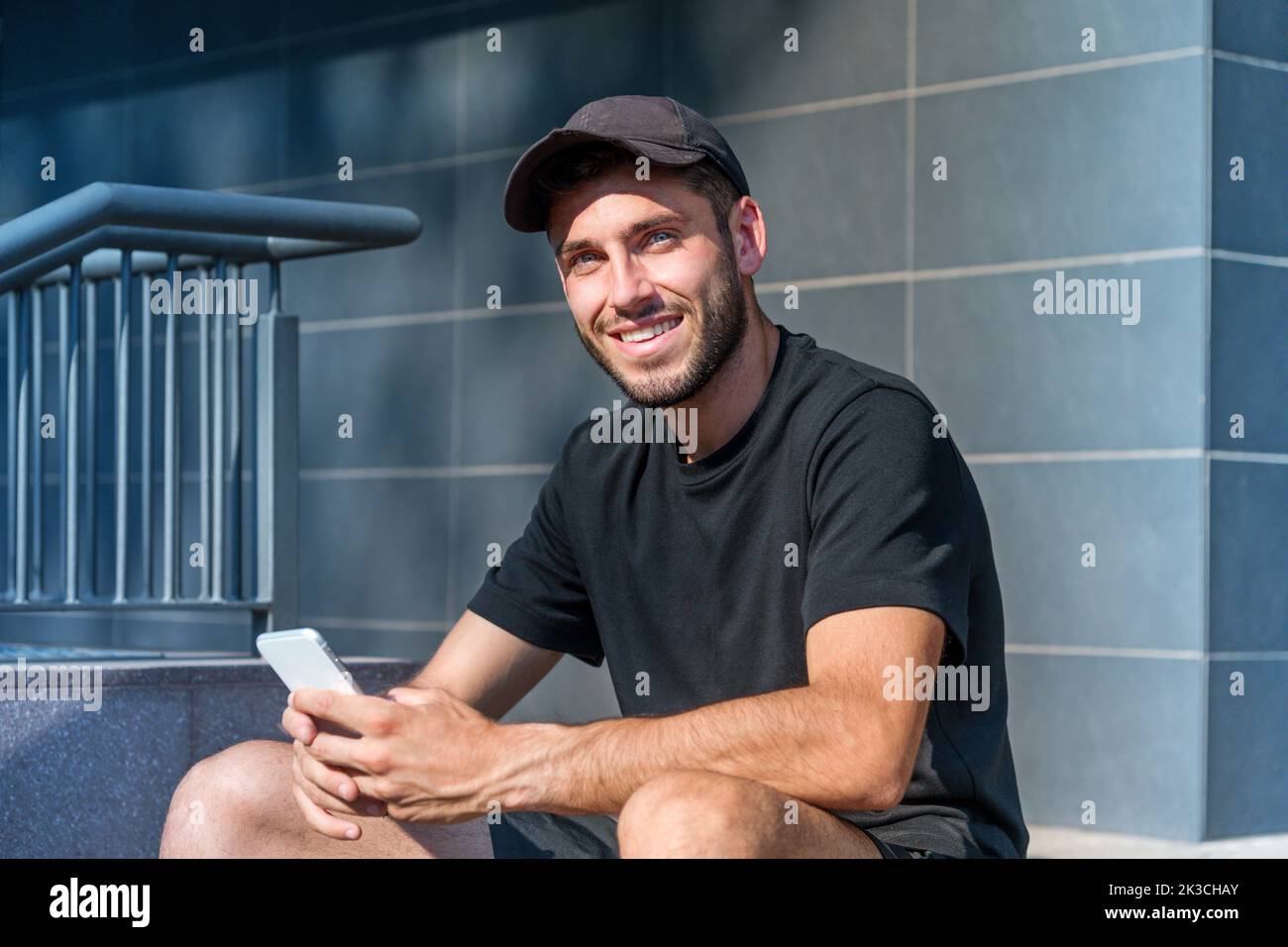 Fröhlicher junger Mann in schwarzem T-Shirt und Hut, der durch sein Handy surft und mit einem Lächeln auf die Kamera blickt, während er auf einer Treppe vor dem modernen Gebäude sitzt Stockfoto