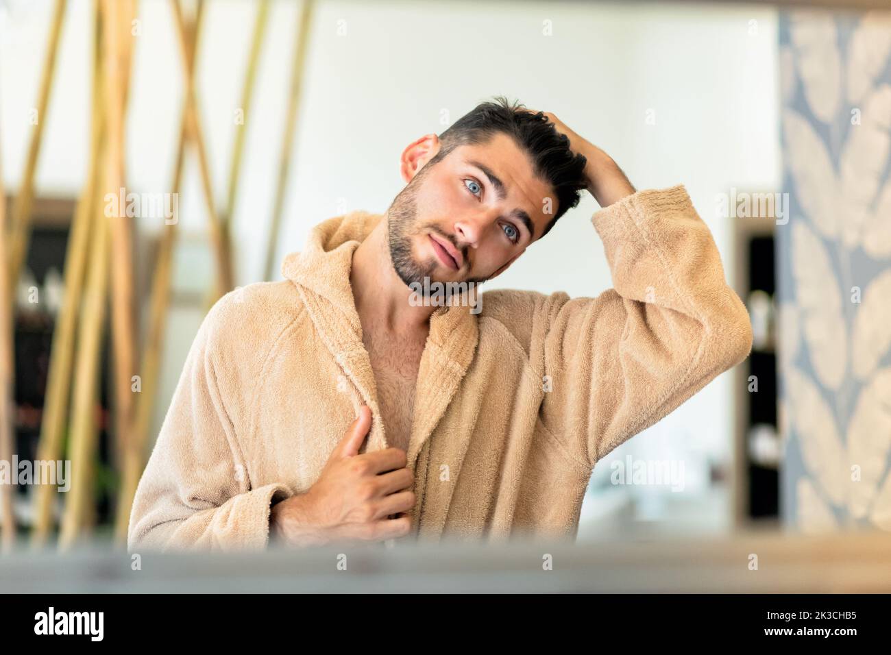 Spiegelung eines jungen Mannes im weichen Bademantel, der während der Spa-Sitzung im Salon dunkle Haare berührt und in den Spiegel schaut Stockfoto