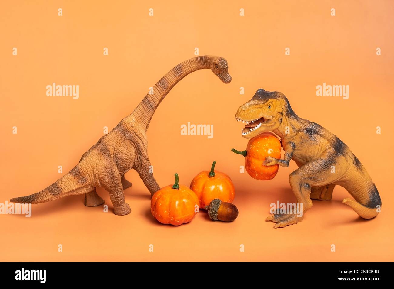 Spielzeug-Dinosaurier Tyrannosaurus hält Kürbis in den Pfoten, auf weißem Podium isoliert auf orangefarbenem Hintergrund Holiday Grußkarte Happy Halloween day, Hello Autumn Creative minimal concept. Stockfoto