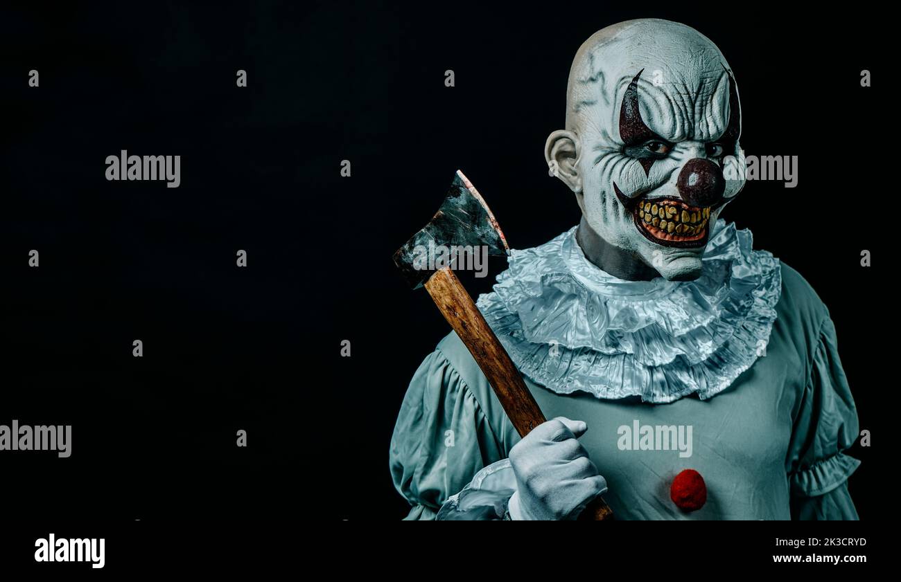 Nahaufnahme eines gruseligen kahlen bösen Clowns, der eine schmutzige alte Axt mit Blutflecken hält und den Beobachter vor einem schwarzen Hintergrund mit einigen leeren s anstarrt Stockfoto