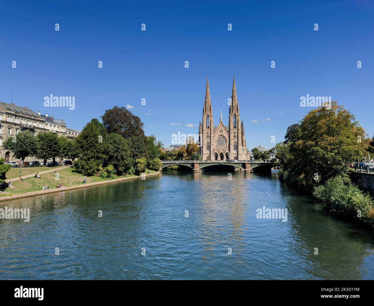 Straßburg, Frankreich - 21. Sep 2022: Postkartenansicht der St. Pauls Kirche und des großen Flusses Ill im Stadtzentrum - klarer blauer Himmel mit einigen Wolken - Menschen, die auf grünem Gras ruhen und die Aussicht bewundern Stockfoto