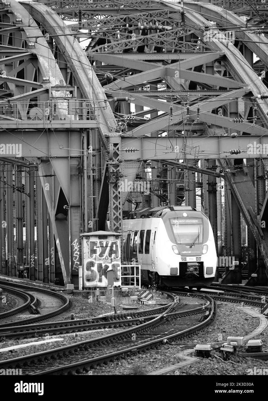 Eine vertikale Graustufenaufnahme eines ankommenden Zuges in der Nähe des Hauptbahnhofs, Köln, Deutschland, über die Hohenzollern Brücke Stockfoto