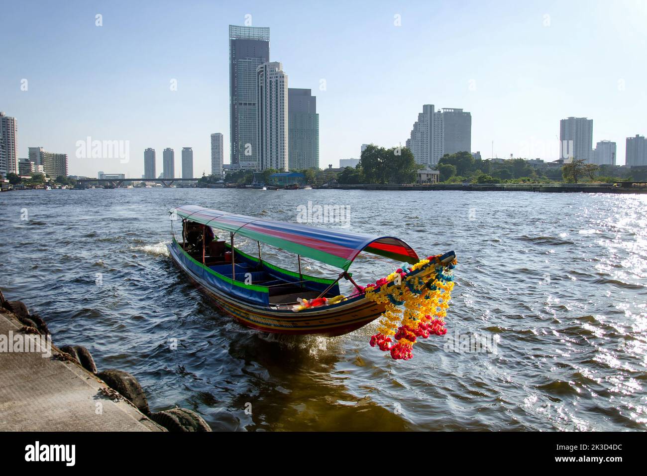 Traditionelles buntes Langboot, das auf dem Chao Phraya Fluss segelt, umgeben von modernen Wolkenkratzern im Hintergrund. Stockfoto