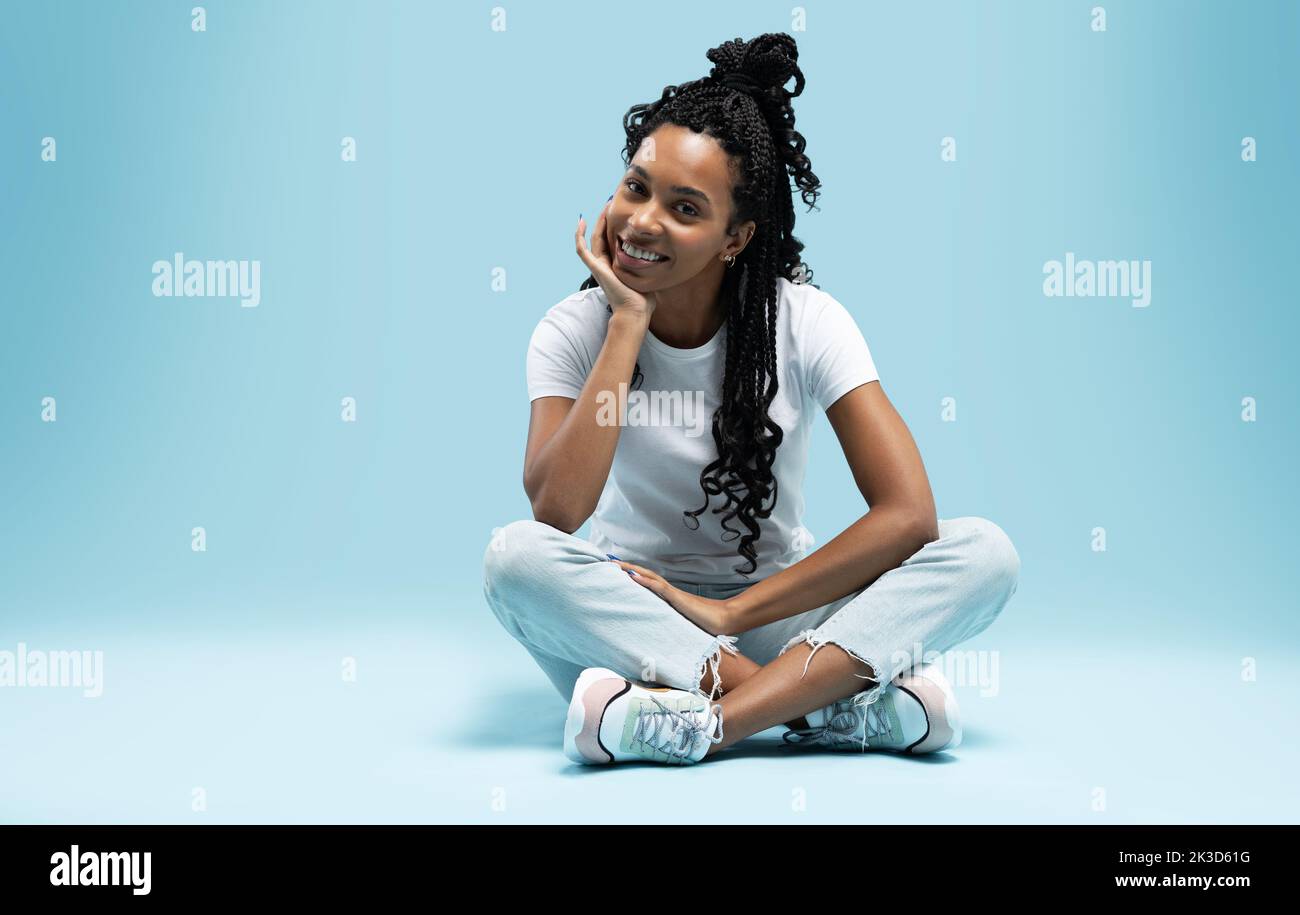 Porträt einer lächelnden jungen afroamerikanischen Frau, die sich ausruhte, während sie auf einem Boden saß, wobei die Beine isoliert auf blauem Hintergrund gekreuzt wurden Stockfoto