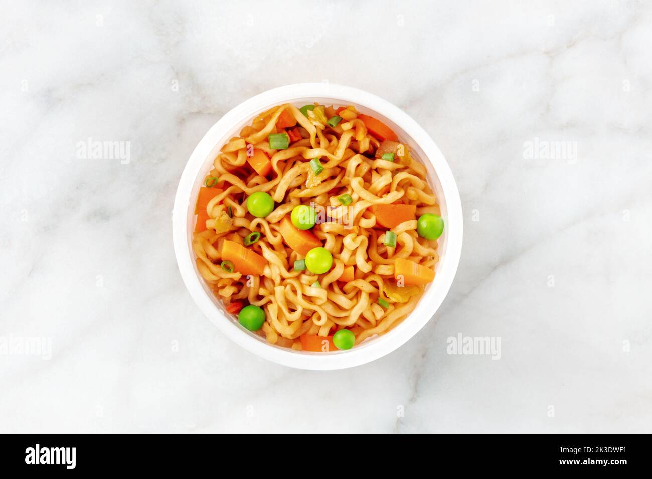 Ramen-Tasse, Instant-Soba-Nudeln in einem Plastikbecher mit Gemüse, grünen Zwiebeln, Erbsen und Karotten, über Kopf auf weißem Marmorboden geschossen Stockfoto