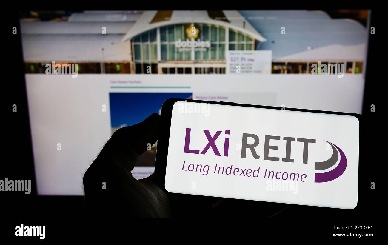 Person, die Mobiltelefon mit dem Logo der britischen Immobiliengesellschaft LXi REIT auf dem Bildschirm vor der Business-Webseite hält. Konzentrieren Sie sich auf die Telefonanzeige. Stockfoto