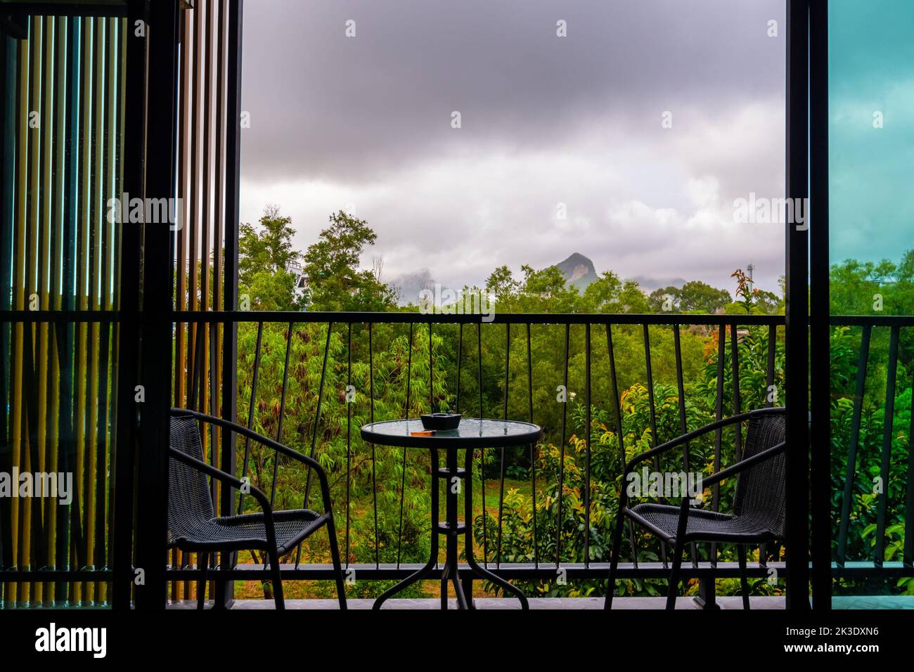 Blick vom Fenster des Hotelzimmers mit Balkon auf den tropischen Wald während des Regensturms. Große Schattenwolken und regnerisches Wetter. Zwei Stühle an der Seite wi Stockfoto