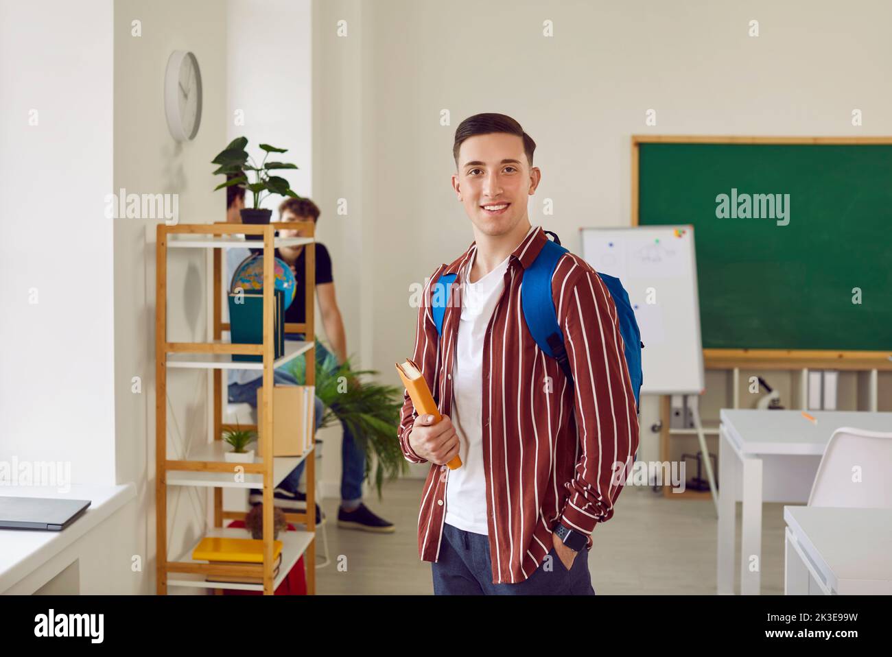 Student Kerl in gestreiftem Hemd mit Rucksack und Buch im Klassenzimmer lächelnd und Blick auf die Kamera. Stockfoto
