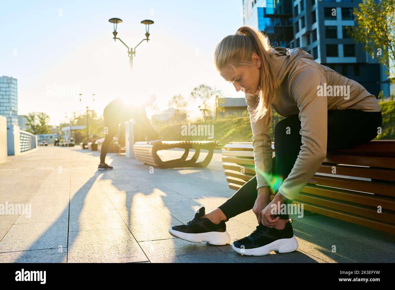 Junge blonde Sportlerin, die sich am Morgen auf einer Holzbank in städtischer Umgebung über das Knie beugte Stockfoto