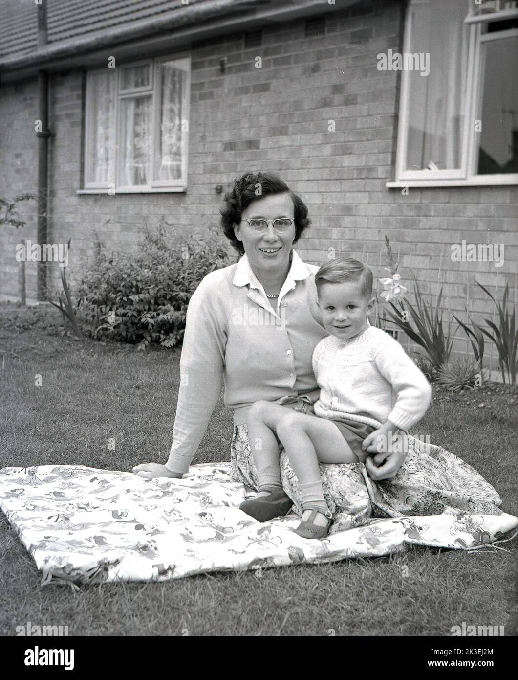 1962, historisch, saß eine Mutter, die vor einem Haus auf einem gemusterten Kinderteppich saß, mit ihrem kleinen Sohn auf ihrem Schoß, Hull, England. Stockfoto