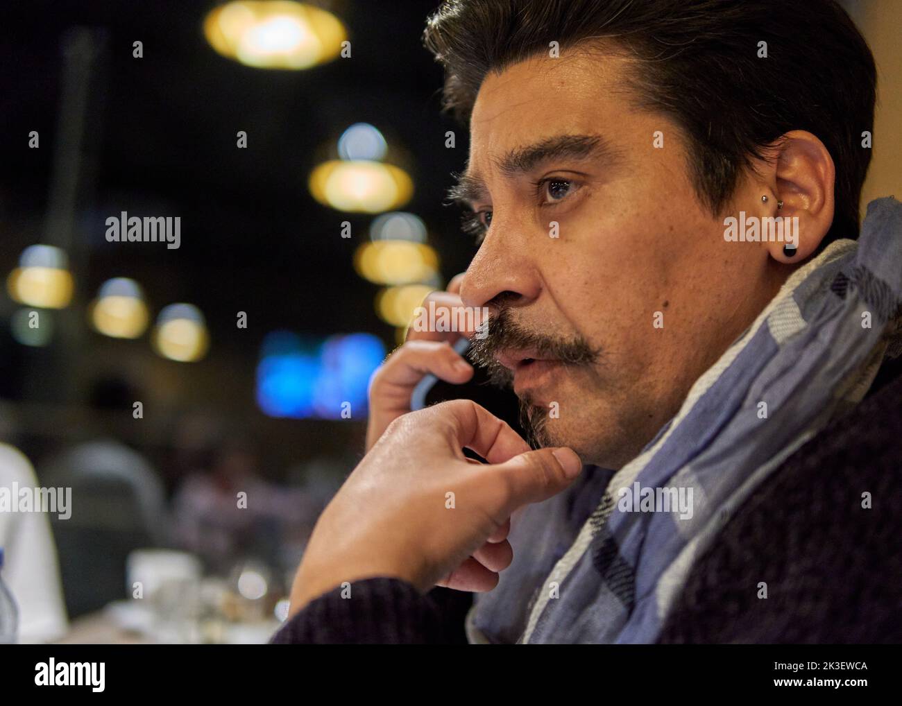 Lateinischer Brünette Mann mit Schnurrbart im Profil sitzt in einer Bar im Gespräch auf dem Handy. Horizontal. Selektiver Fokus und Hintergrundbeleuchtung Stockfoto