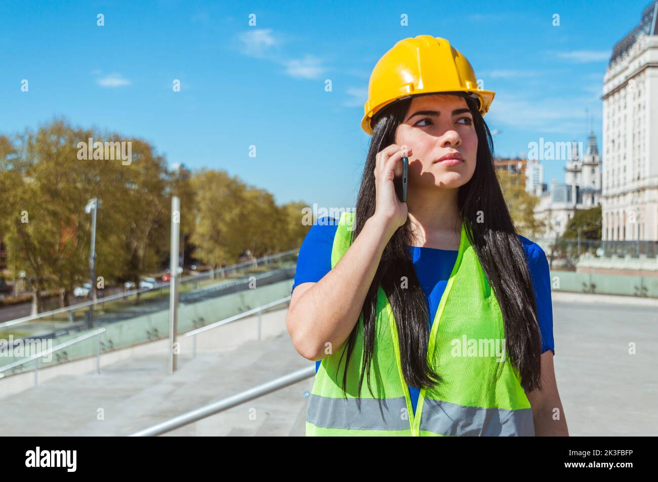 lateinisch kaukasische junge venezolanische Frau, Ingenieurin mit gelbem Helm, Weste und blauer Kleidung, im Freien mit Zweifeln, die mit ihrem Chef am Telefon sprechen, Tech Stockfoto