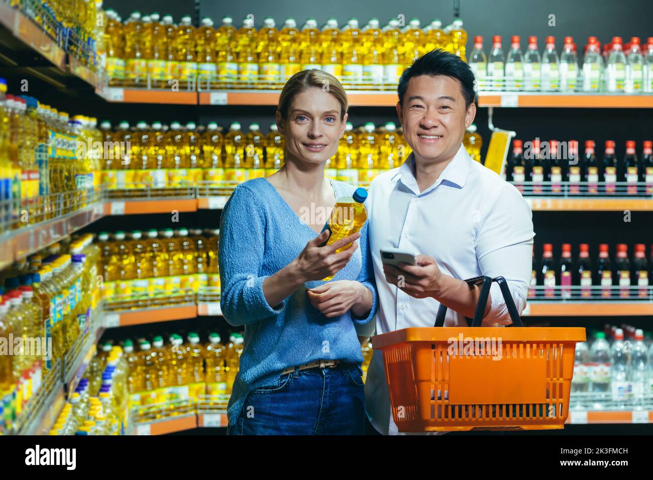 Ein junges internationales Paar, eine Frau und ein asiatischer Mann, entscheiden sich in einem Supermarkt für Öl. Sie halten eine Flasche Öl, einen Korb und ein Telefon in der Hand. Sie schauen auf die Kamera und lächeln. Stockfoto