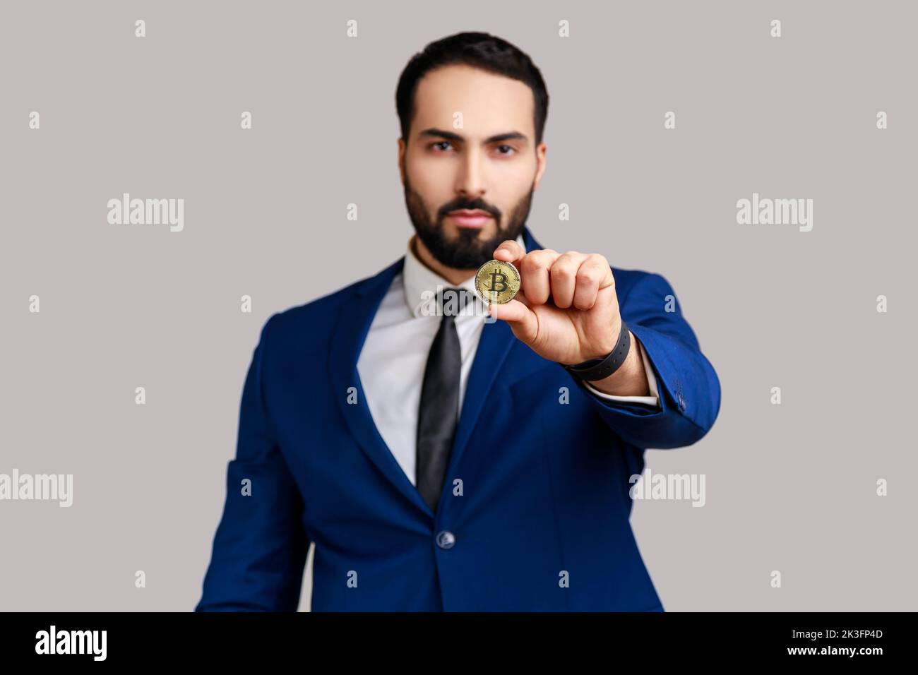 Ein hübscher bärtiger Mann, der Goldbitcoin aushält, die Kamera mit einem toothy Lächeln anschaut, ein Blockchain-Konzept und einen offiziellen Style-Anzug trägt. Innenaufnahme des Studios isoliert auf grauem Hintergrund. Stockfoto