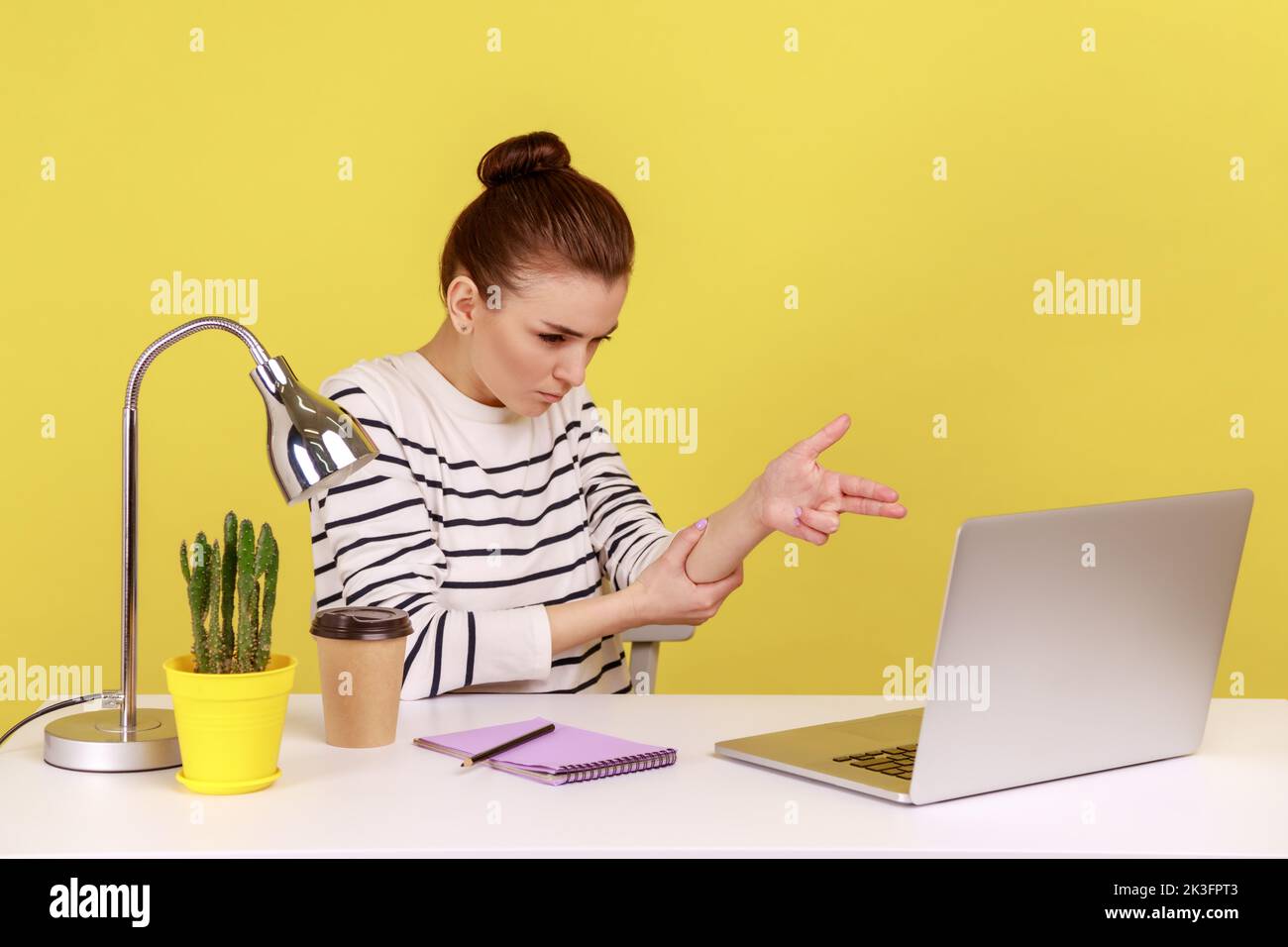 Aggressive Managerin, die am Arbeitsplatz sitzt, mit einem Laptop bedroht und mit dem Finger auf den Laptop-Bildschirm zeigt und mit wütendem Ausdruck schießt. Studio-Innenaufnahme isoliert auf gelbem Hintergrund. Stockfoto