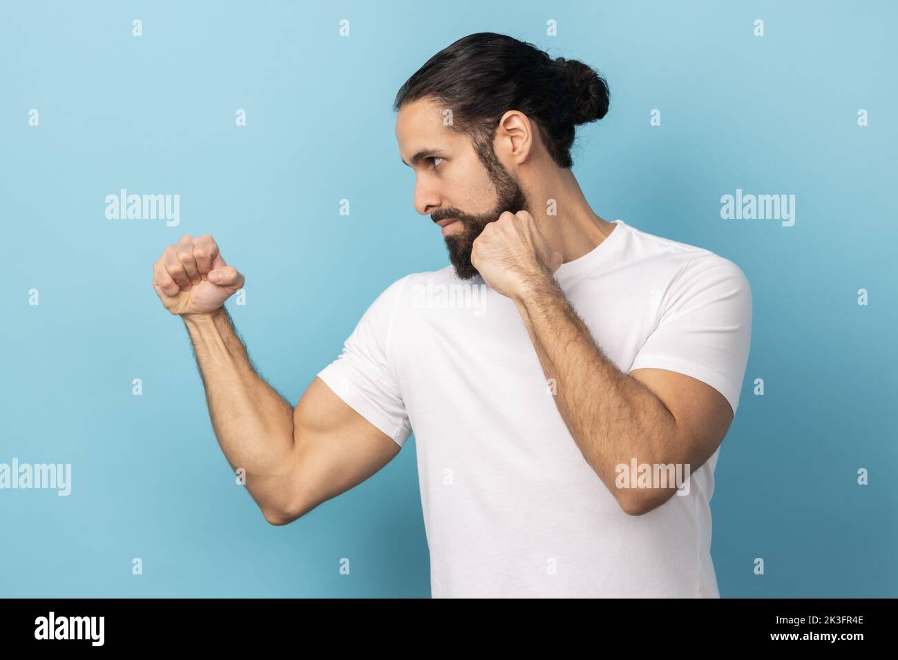Seitenansicht eines aggressiven Mannes mit Bart und weißem T-Shirt, der geballte Fäuste zum Boxen bereithält, Kampfkunsttrainer, Selbstverteidigung. Innenaufnahme des Studios isoliert auf blauem Hintergrund. Stockfoto