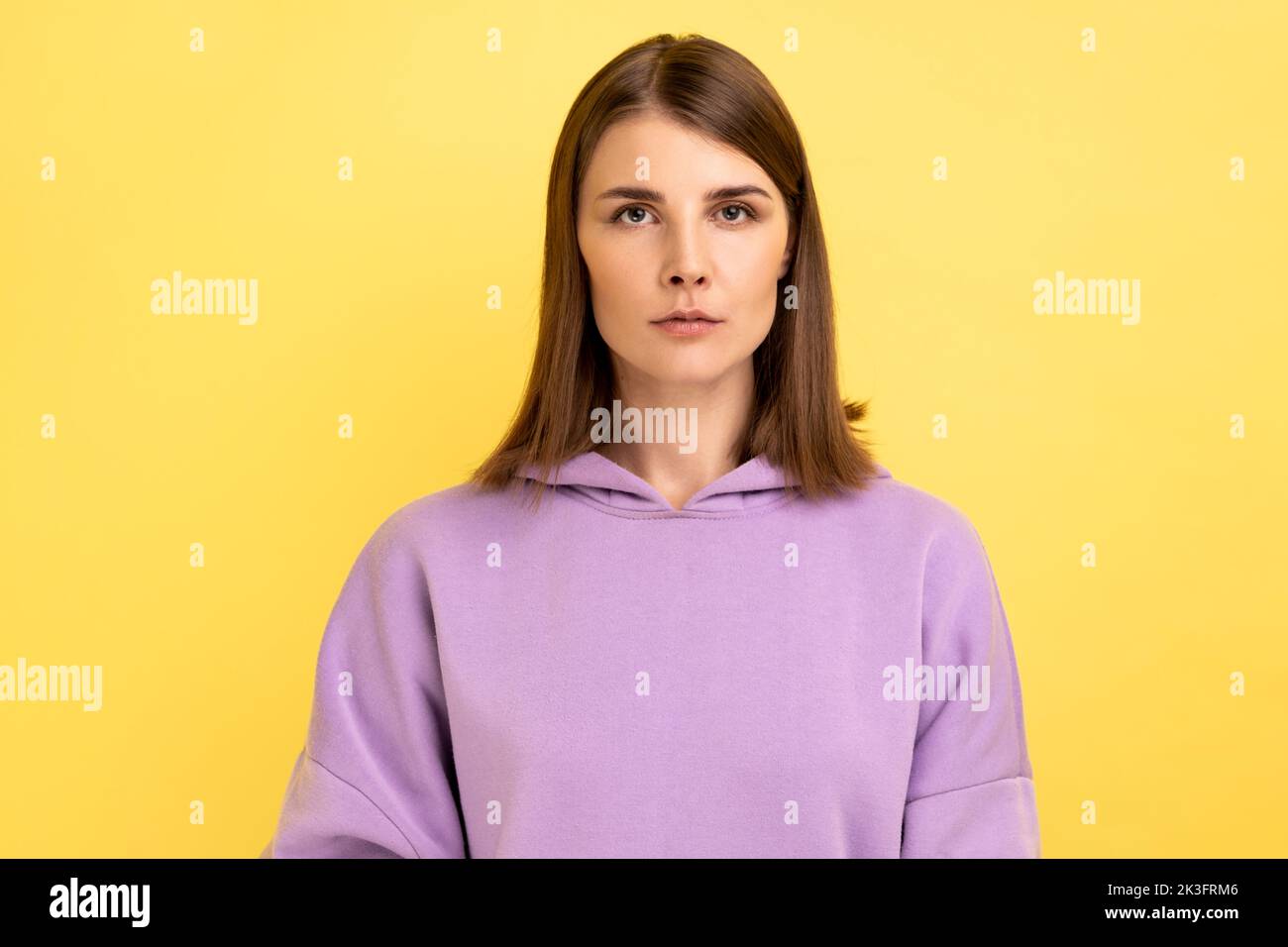 Porträt einer jungen, ernsten, unlächelnden, strengen Frau, die mit einem ruhigen, aufmerksamen Gesichtsausdruck auf die Kamera blickt und einen purpurnen Hoodie trägt. Innenaufnahme des Studios isoliert auf gelbem Hintergrund. Stockfoto