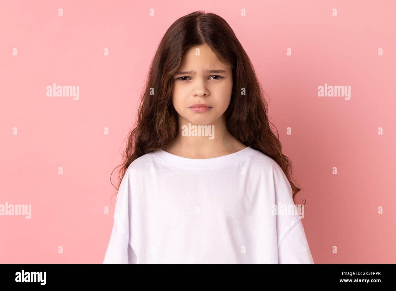 Portrait eines verärgerten kleinen Mädchens, das weißes T-Shirt trägt, das negative Emotionen und Trauer ausdrückt, schlechte Laune hat, traurig und unglücklich ist. Innenaufnahme des Studios isoliert auf rosa Hintergrund. Stockfoto