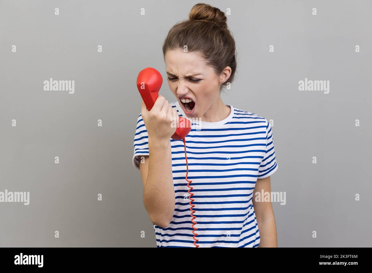 Porträt einer wütenden, nervösen Frau, die gestreiftes T-Shirt trägt und schreiend im Retro-Festnetztelefon redet und sich über die Verbindungsqualität beschwert. Innenaufnahme des Studios isoliert auf grauem Hintergrund. Stockfoto