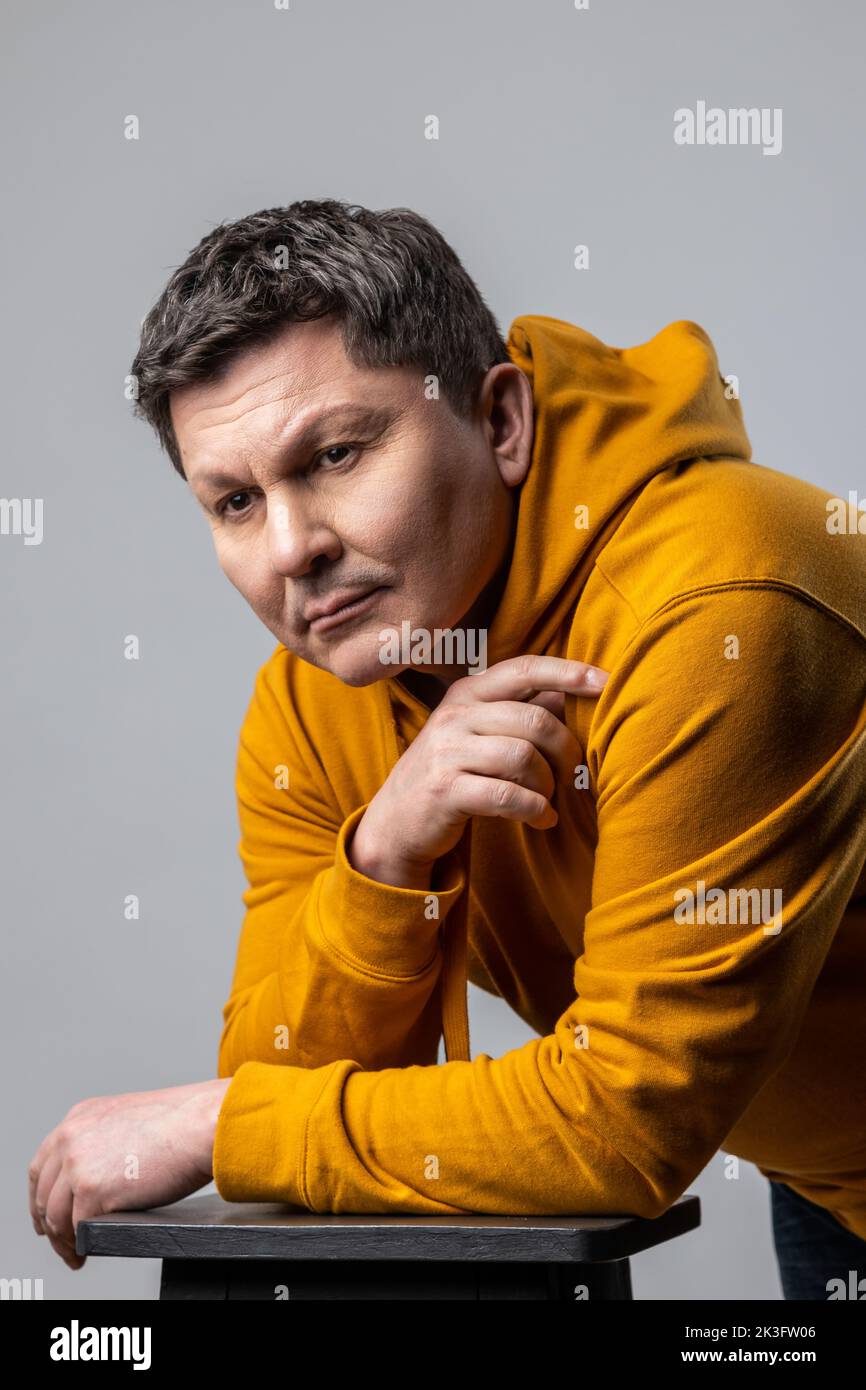 Porträt eines gutaussehenden Mannes mittleren Alters, der nachdenklich aussieht, sich auf einen Stuhl lehnt, tief in Gedanken ist und einen Hoodie im urbanen Stil trägt. Innenaufnahme des Studios isoliert auf grauem Hintergrund. Stockfoto