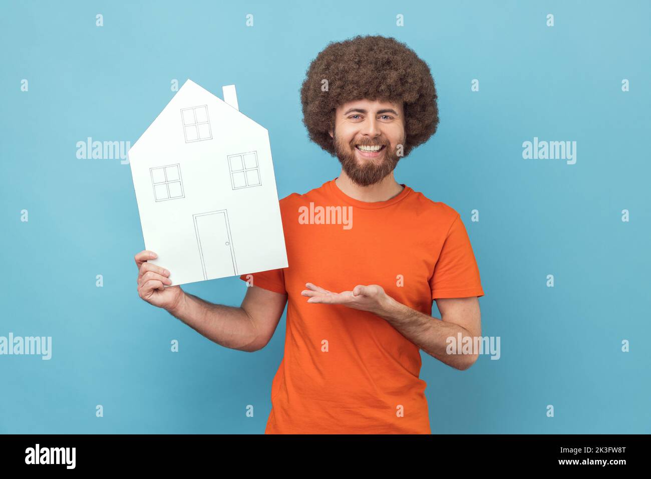 Mann mit Afro-Frisur präsentiert Papierhaus mit gezogenen Fenstern und Türen, macht Projekt von zu Hause aus ihren Träumen, Blick auf die Kamera mit einem Lächeln. Innenaufnahme des Studios isoliert auf blauem Hintergrund. Stockfoto