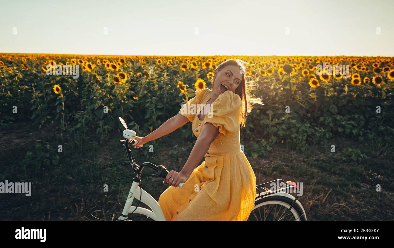 Ländliche Frau in zeitlosem Kleid Reiten Retro-Stil weißen Fahrrad auf der Landstraße allein in der Nähe Sonnenblumen Feld. Vintage-Mode, tolles Abenteuer Stockfoto