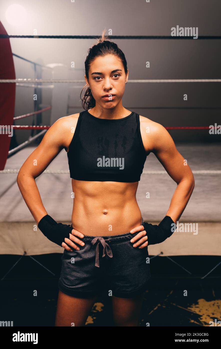 Selbstbewusste Boxerin, die die Kamera anschaut, während sie vor einem Boxring im Fitnessstudio steht. Athletische junge Frau, die in einer Boxhalle trainiert. Stockfoto