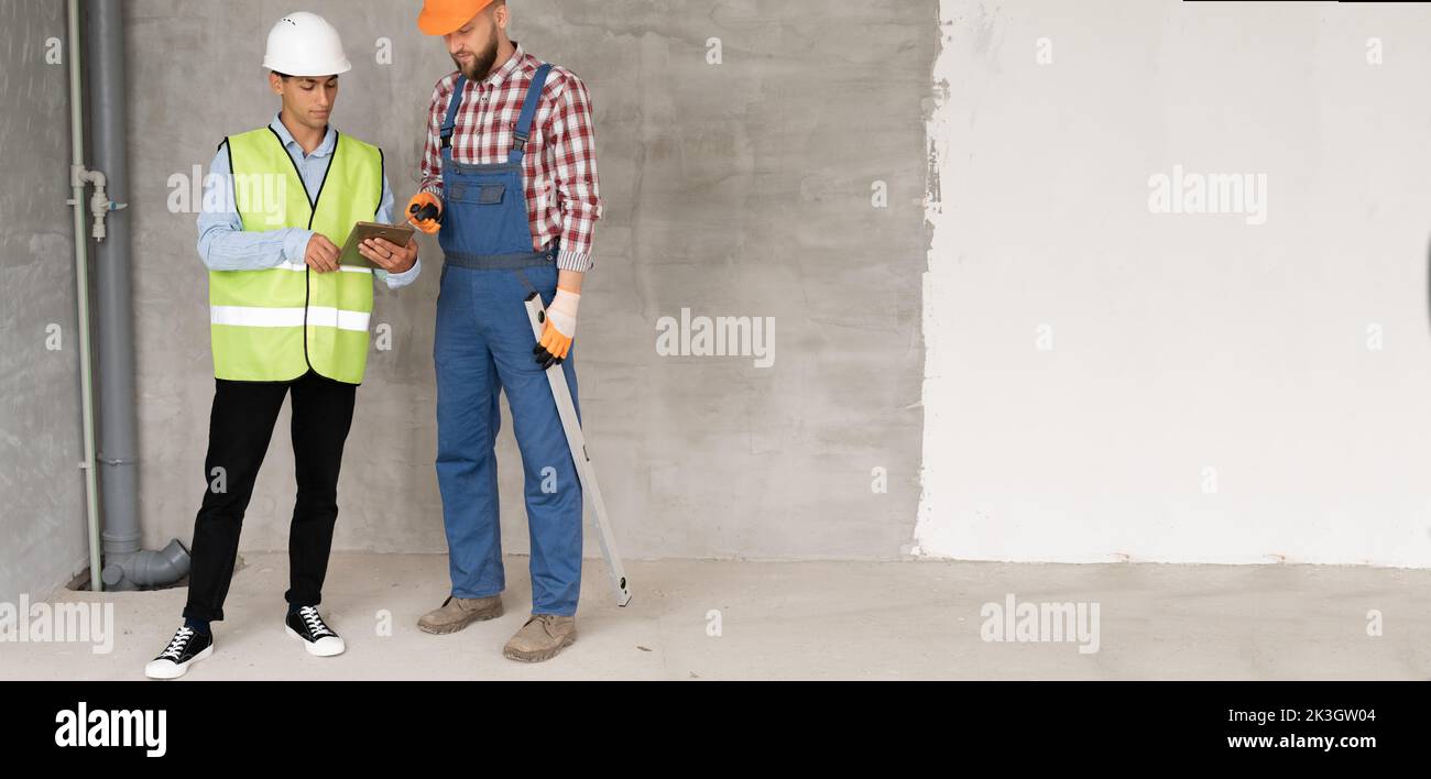 Zwei männliche Baumeister und Ingenieur in Schutzhelmen arbeiten auf einer Baustelle mit einem Tablet und diskutieren Baupläne für die Renovierung von Wohnheimen. Stockfoto