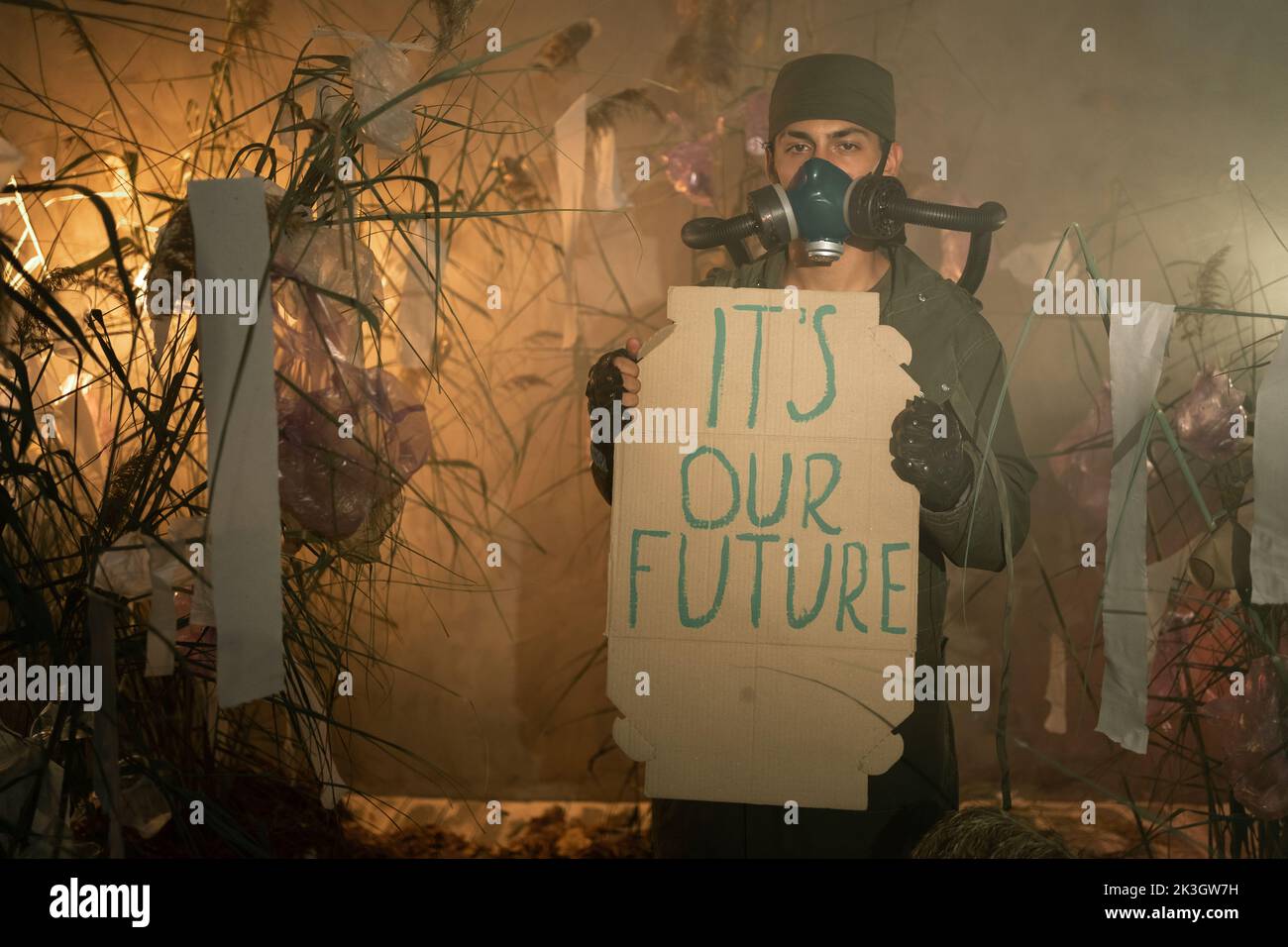 Ein Mann in einer Gasmaske hält Plakat mit Worten seine unsere Zukunft. Brennender Plastikmüll auf dem Hintergrund. Umweltprobleme: Plastikmüll Stockfoto