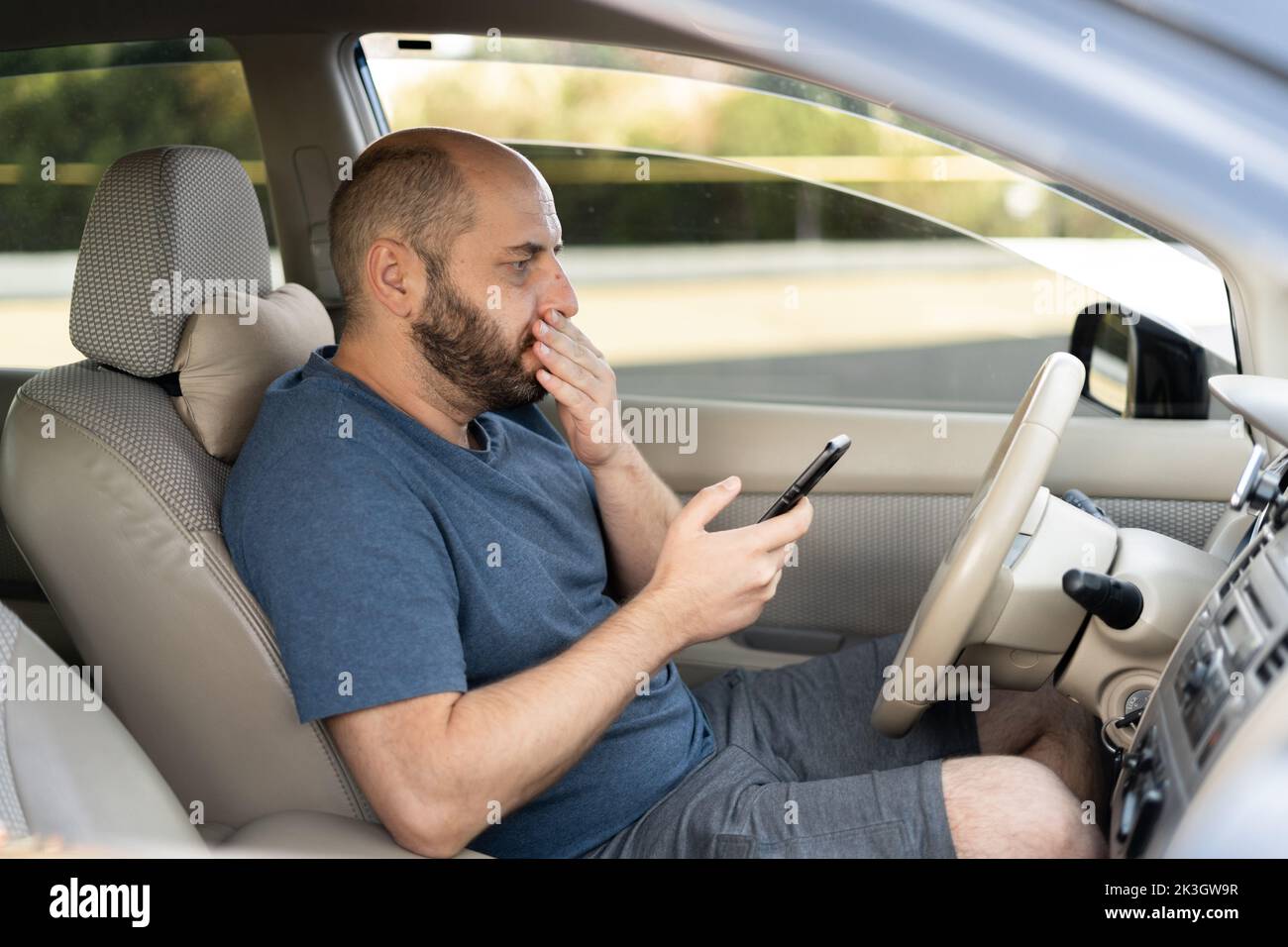 Mann sitzt im Auto mit Handy in der Hand SMS. Schockierter Mann, der sein Smartphone überprüft und auf der Straße nicht aufpasst, betäubt durch schlechte SMS Stockfoto