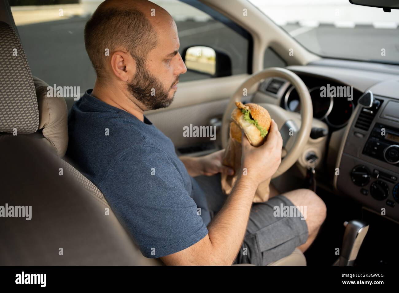 Mann, der einen Hamburger isst, während er mit dem Auto fährt. Hungriger Fahrer, der sein Auto fährt, während er im Verkehr Essen isst Stockfoto