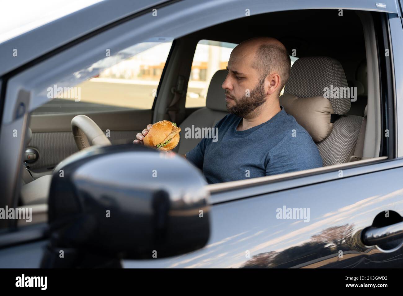 Fahrer Mann am Steuer beim Essen im Verkehr, essen Hamburger im Auto. Stadtleben. Stockfoto