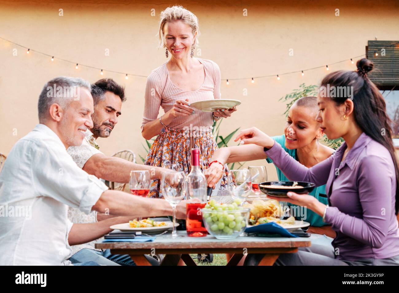 Reife Frau, die Freunden Essen serviert und Spaß bei einem Grilltreffen hat. Eine Gruppe von erwachsenen Freunden, die im Hinterhof eine Dinnerparty im Freien machen Stockfoto