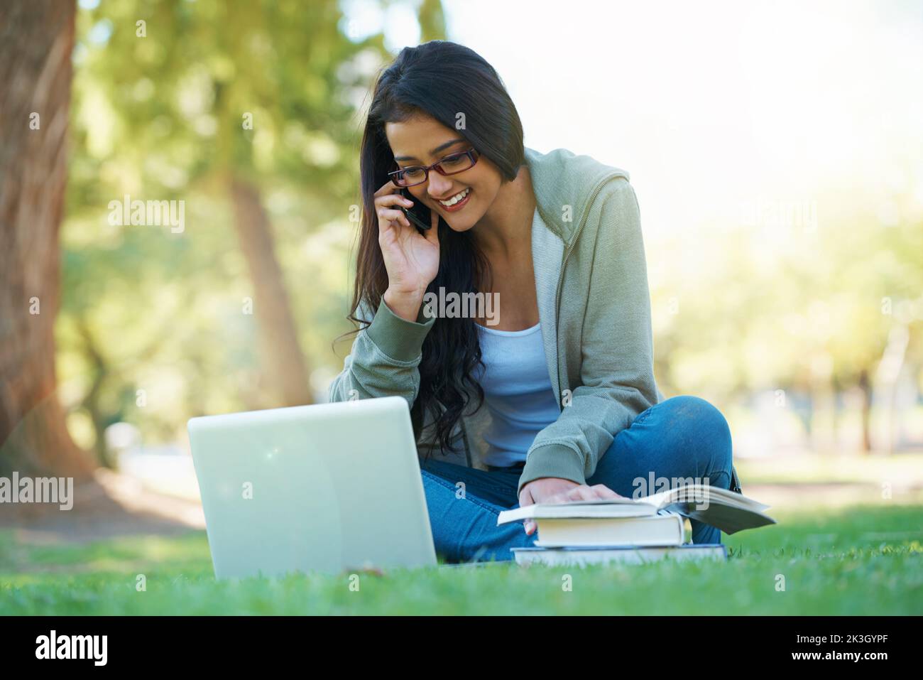 Treffen Sie mich im Park zu einer Studiensitzung. Eine junge Studentin spricht auf ihrer Zelle, während sie mit einem Laptop und Testbüchern im Park sitzt. Stockfoto