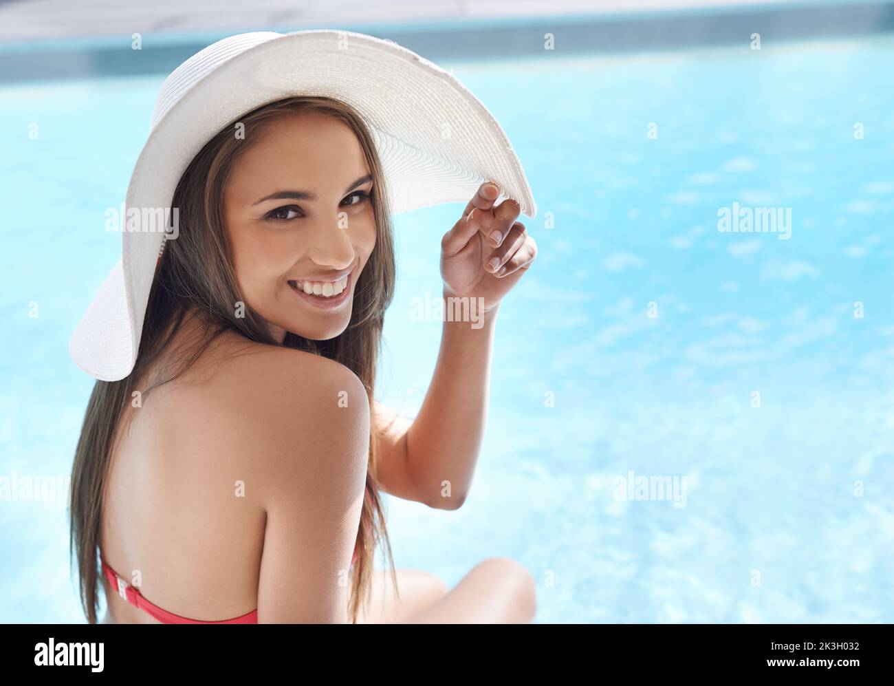 Der Pool ist der einzige Ort, an dem man sein sollte. Porträt einer lächelnden jungen Frau, die mit einem Sonnenhut an einem Pool sitzt. Stockfoto