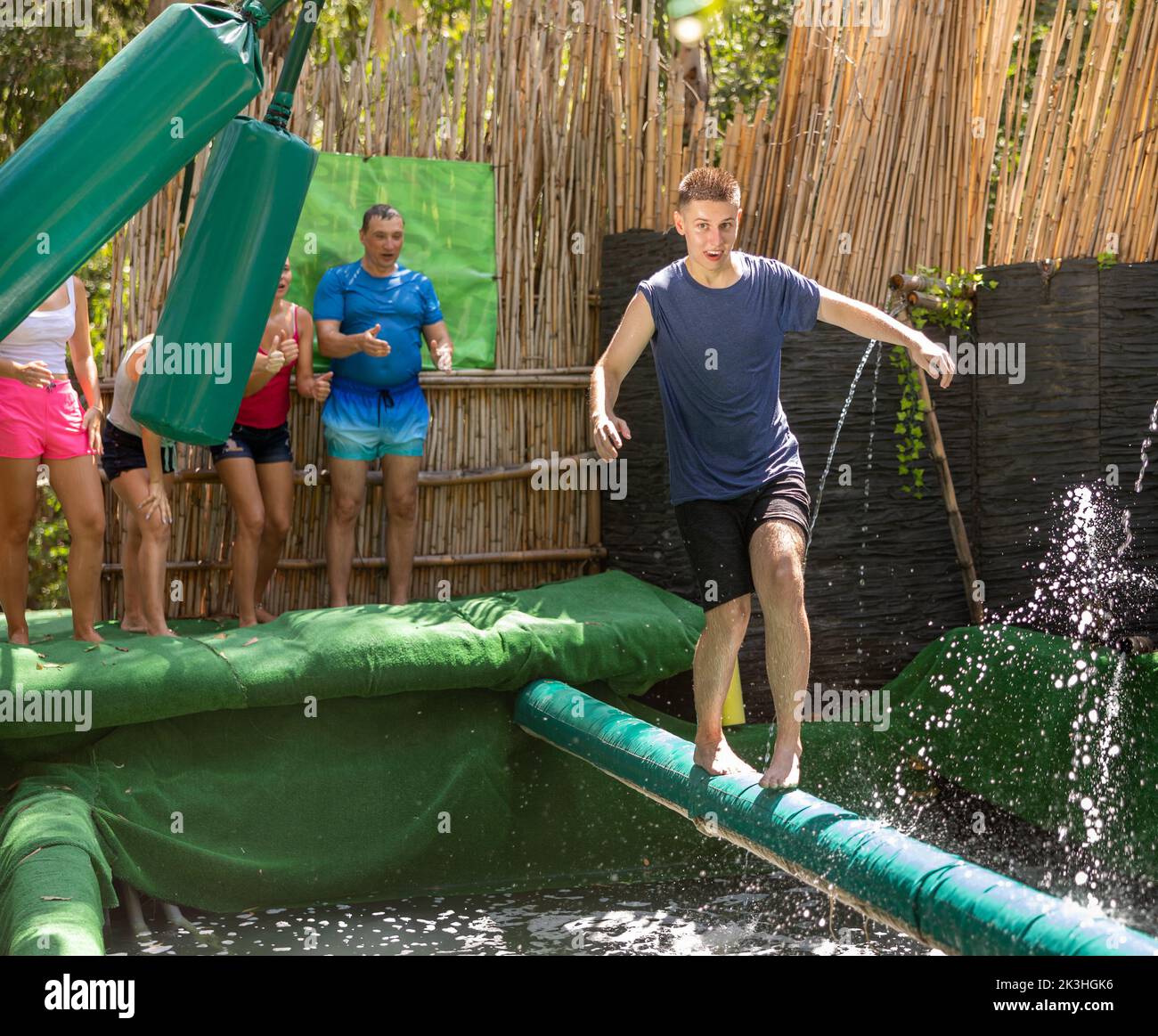 Fröhlicher Kerl balanciert auf weichem Holz zwischen schaukelnden Taschen über Pool mit Wasser Stockfoto