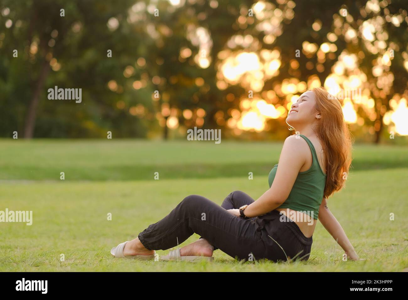 Junge Asiatin mit langen karamellfarbenen, welligen Haaren, grünem Tank-Top und schwarzer Leinenhose sitzt auf dem Gras und sonnt sich bei Sonnenuntergang in der Wärme der Sonne. Stockfoto