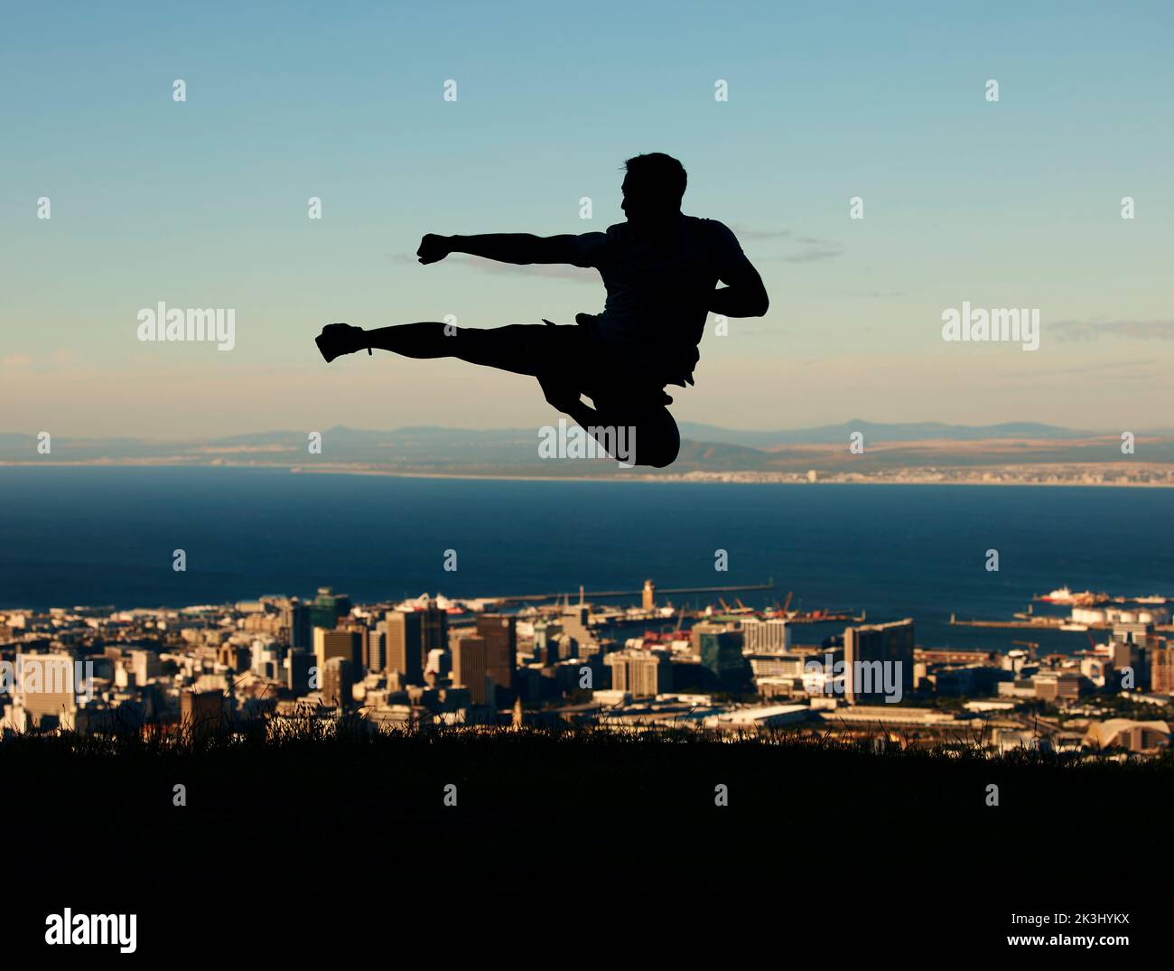 Silhouette eines Mannes, der Karate mit Himmel und Stadt im Hintergrund macht. Umriss eines männlichen Athleten, der in der Kampfkunst-Bewegung in die Luft tritt und tritt Stockfoto