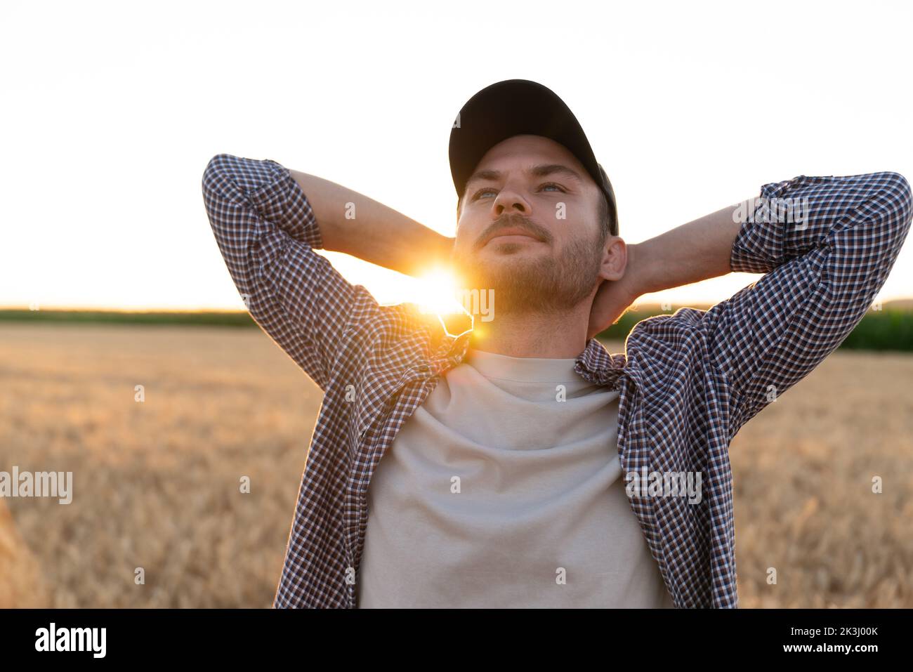 Bärtiger Bauer mit den Händen hinter dem Kopf auf dem landwirtschaftlichen Feld bei Sonnenuntergang Stockfoto