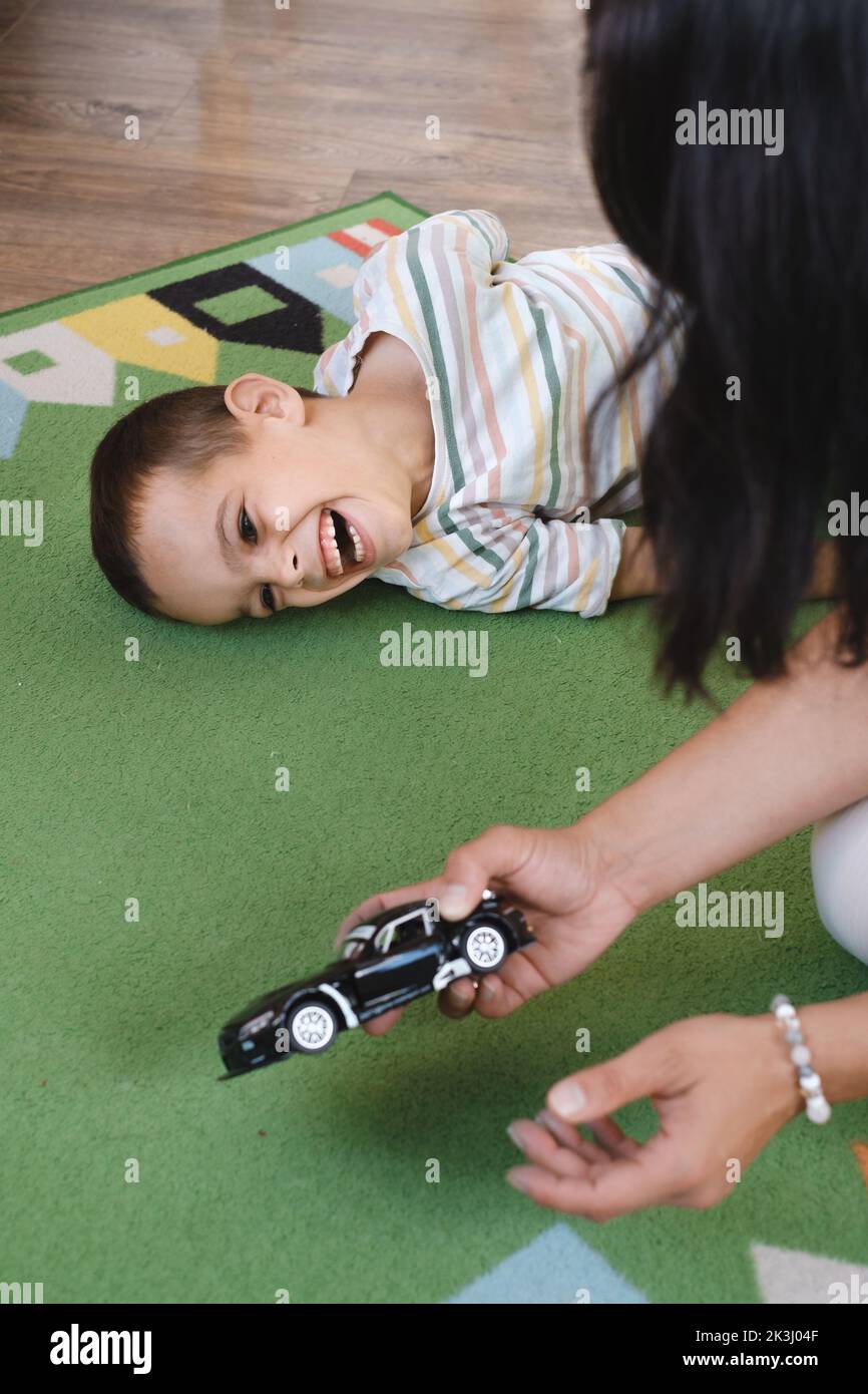 Behinderter Junge spielt Spielzeugautos mit Mutter zu Hause. Zerebrale Lähmung Kind unterhaltsam auf der Matte mit Betreuer. Kommunikation und Rehabilitation in Stockfoto