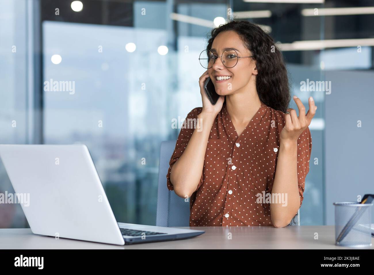 Junge, erfolgreiche hispanische Geschäftsfrau, die im modernen Büro arbeitet, weibliche Angestellte, die fröhlich mit Kollegen telefoniert und lächelt und am Schreibtisch sitzt, mit einem Laptop. Stockfoto