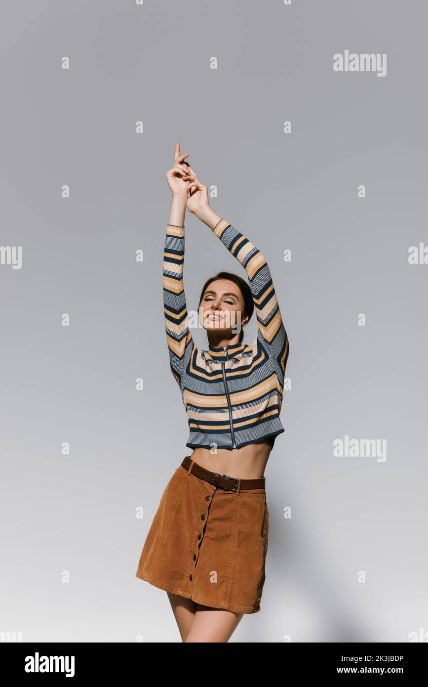 Glückliche junge Frau in gestreiften Rollkragen und Rock posiert mit erhobenen Händen isoliert auf grau, Stock Bild Stockfoto
