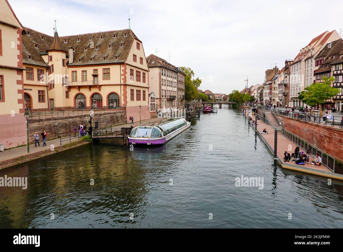 Von der Corbeau-Brücke aus blicken Sie auf einen der Kanäle, während die Menschen sich entspannen, am Quai entlang spazieren und an einem Touristenboot im historischen Straßburg, Frankreich, vorbeifahren. Stockfoto