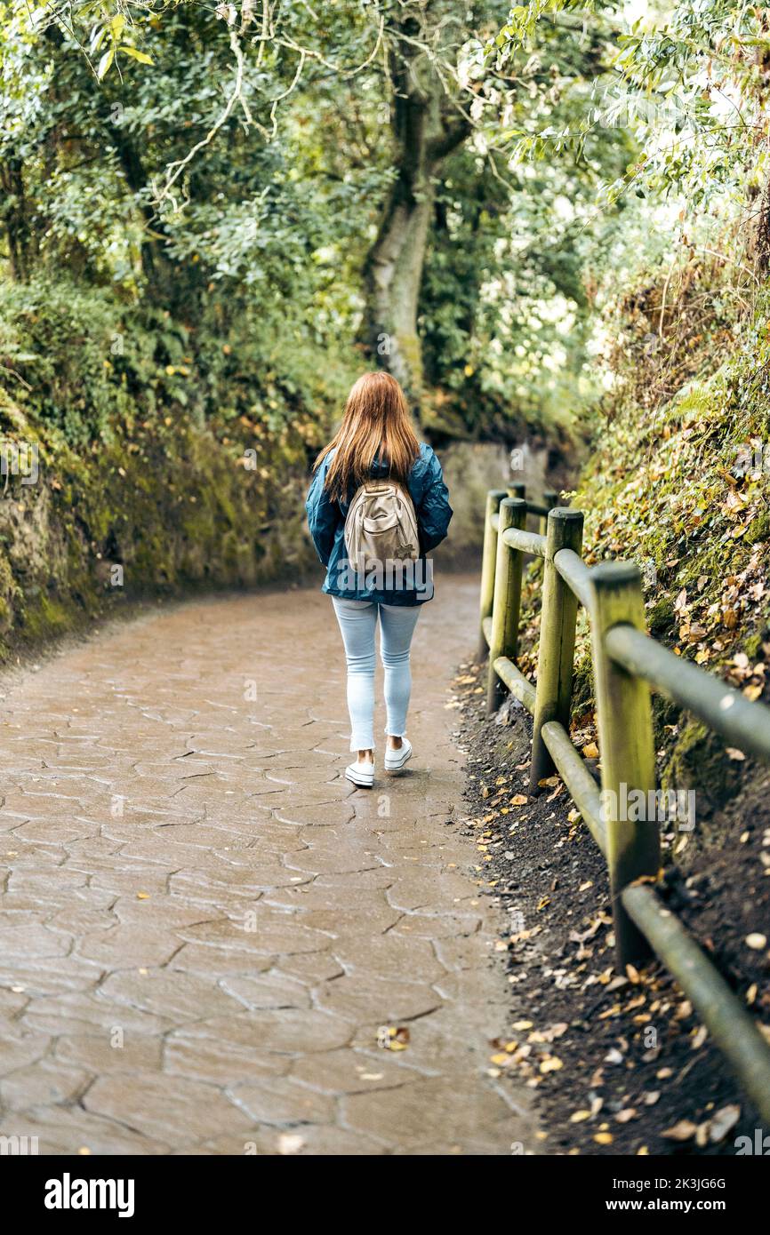 Frau, die mit Regenmantel und Rucksack auf einer gepflasterten Straße unterwegs ist. Stockfoto