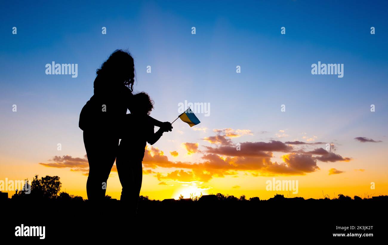 Mutter mit einem Kind vor dem Hintergrund eines blau-gelben Sonnenuntergangshimmels mit der Flagge der ukraine in der Hand, ukrainischer Sieg im Krieg. Stockfoto