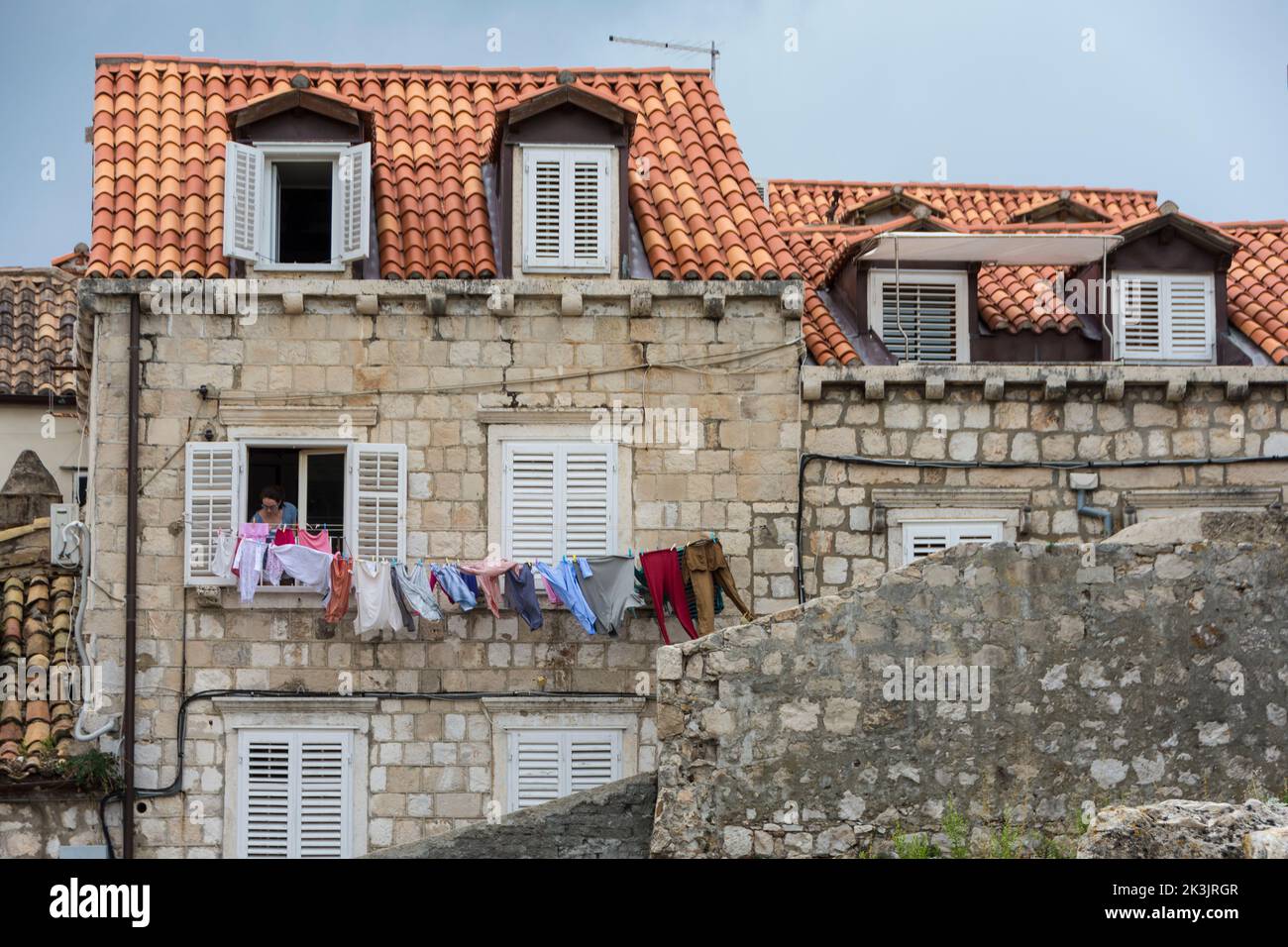 Frau, die die Wäsche an einer Wäscheleine vor einem Fenster mit weißen Fensterläden aufgehängt hat.Altstadt von Dubrovnik, Kroatien Stockfoto