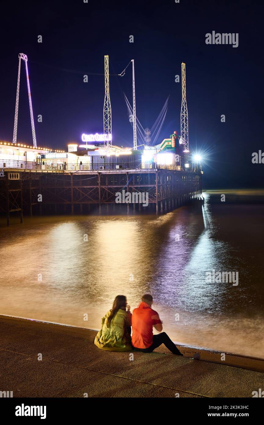 Während der Blackpool-Illuminationen saßen zwei Personen auf der Ufertreppe mit Blick auf den South Pier in der Nacht Stockfoto