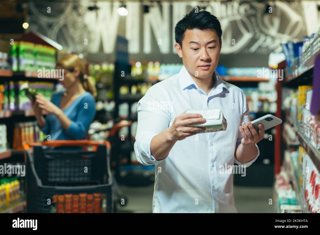 Ein junger asiatischer Mann steht in einem Supermarkt und wählt Pads für seine Frau, Freundin. Er weiß nicht, welchen er wählen soll, er steht nachdenklich, verwirrt und hält das Telefon in der Hand. Stockfoto